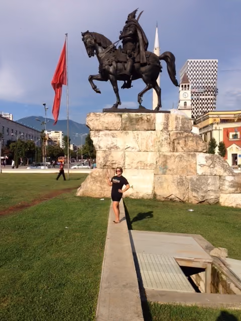 Person posing on a path near a large equestrian statue on a stone pedestal with a red flag nearby.