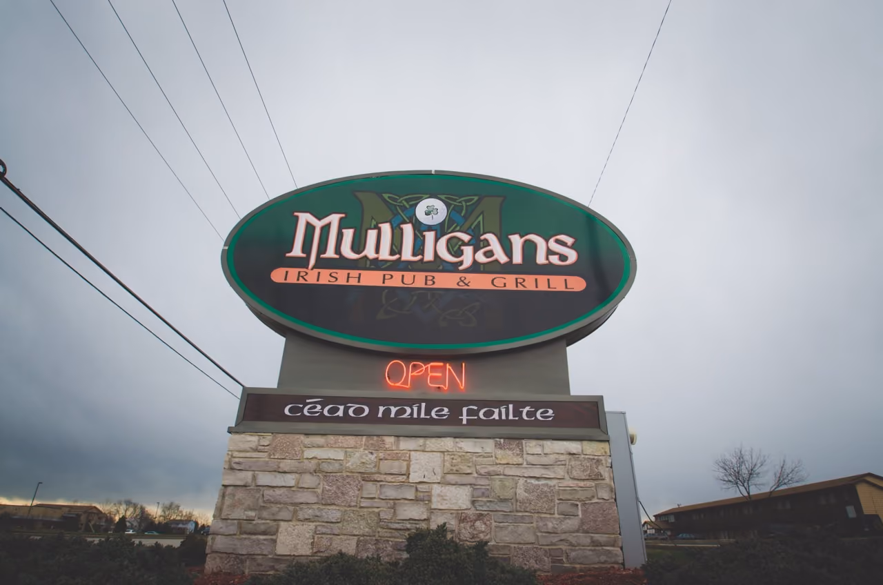 Sign for Mulligans Irish Pub & Grill with neon OPEN and Irish phrase on stone base under overcast sky.