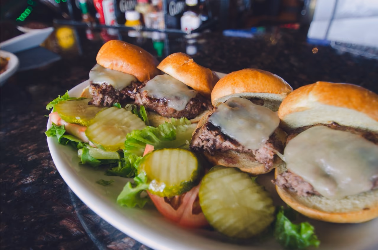Four cheeseburger sliders on buns with lettuce, tomato, and pickle slices on a white plate.