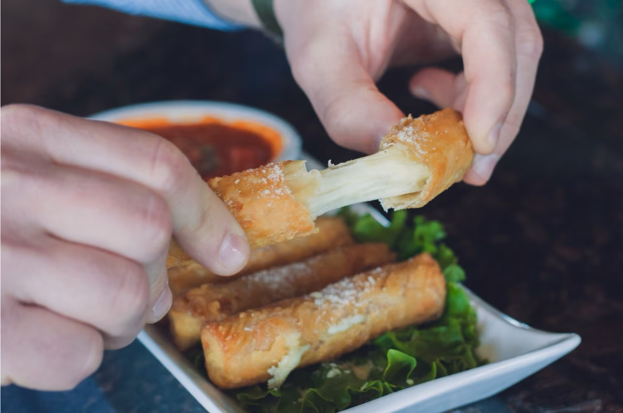 Hands pulling apart a fried mozzarella stick with melted cheese stretching, with sauce and lettuce on plate.