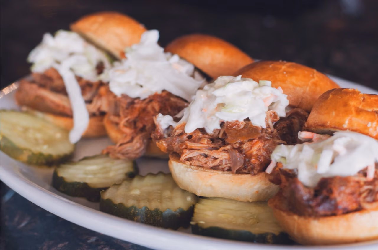 Four pulled pork sliders topped with coleslaw on a white plate with pickle slices.