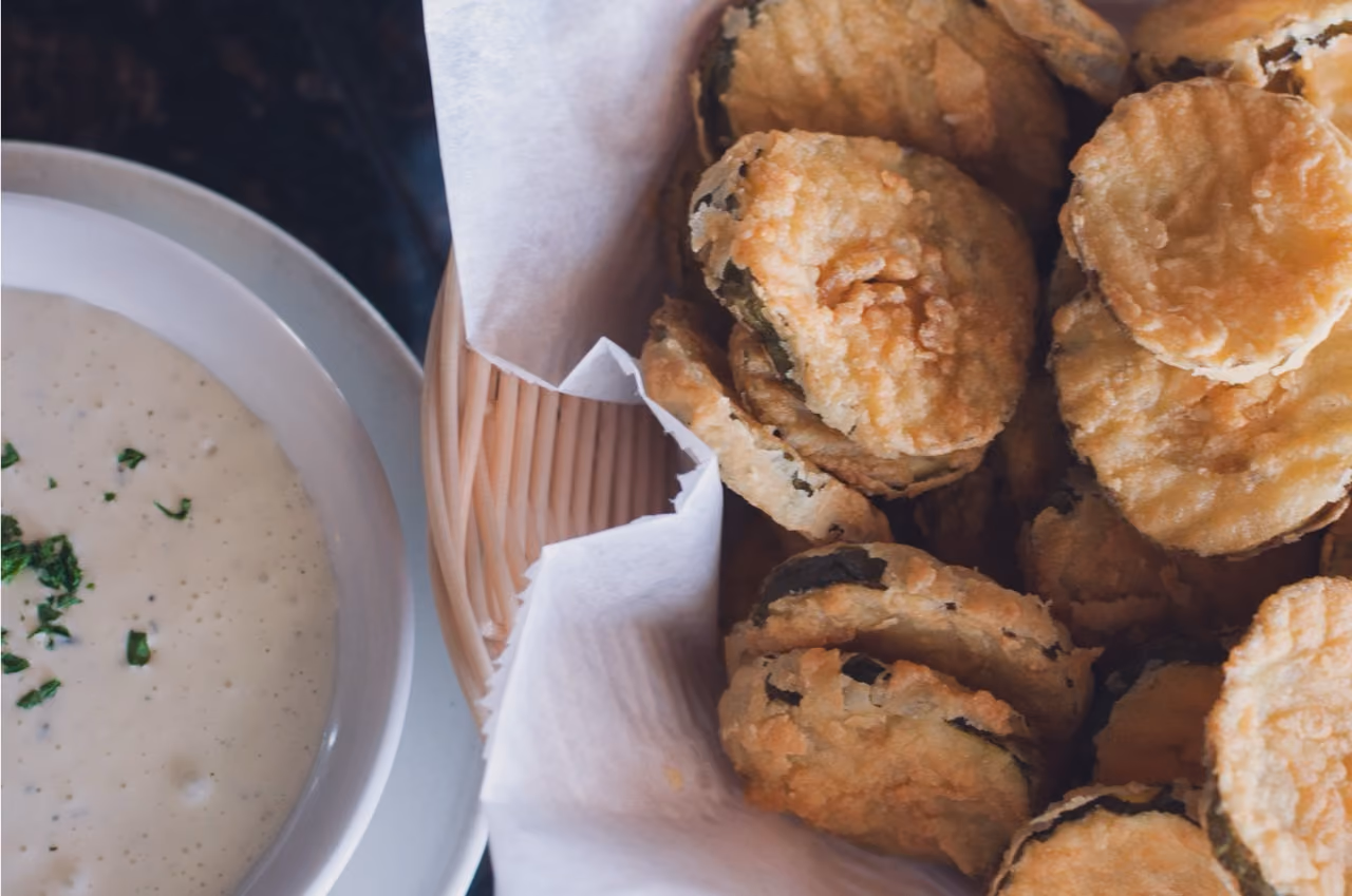 Basket of golden fried pickles with a bowl of creamy dipping sauce garnished with herbs.