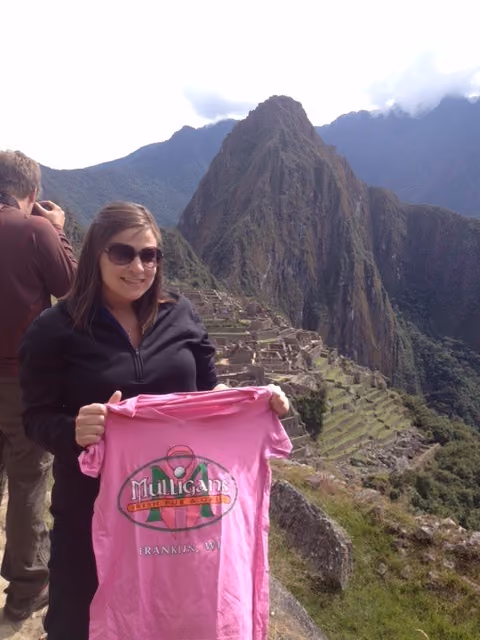 Woman with sunglasses holding a pink Mulligans Irish Pub T-shirt at Machu Picchu ruins.