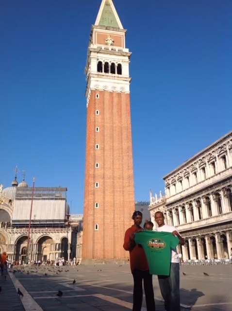 Two men holding a green Mulligans Irish Pub shirt in front of the tall brick bell tower in a city square.