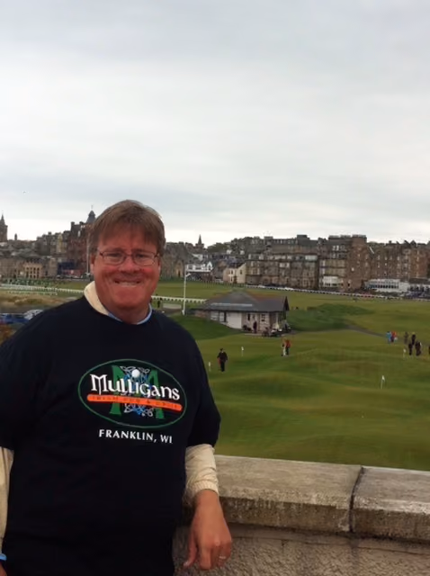 Man smiling wearing a Mulligans Franklin WI shirt with a golf course and buildings in the background.