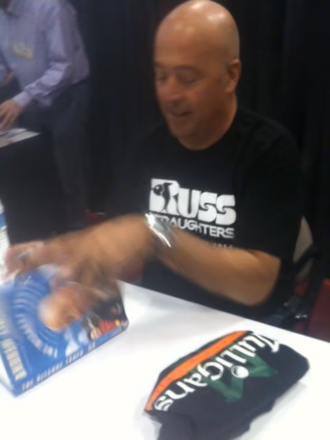 Man in black shirt signing a blue book at a table with folded clothing beside him.