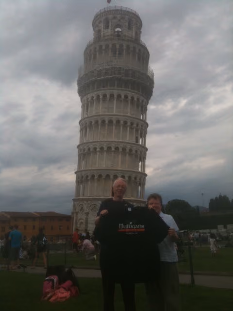 Two people holding a Mulligans Irish Pub shirt in front of the Leaning Tower of Pisa on a cloudy day.