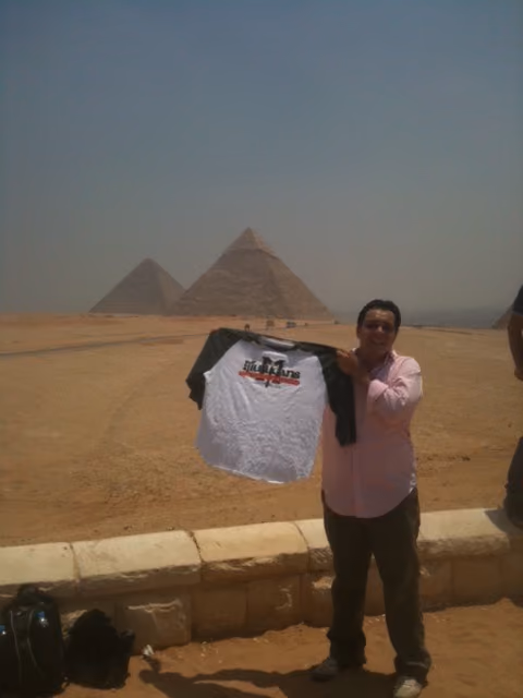 Man in pink shirt holding a white-black shirt with pyramids in the hazy background.
