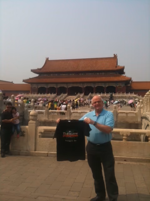 Man holding a Mulligans t-shirt in front of a historic Chinese palace with crowd and stone railings.