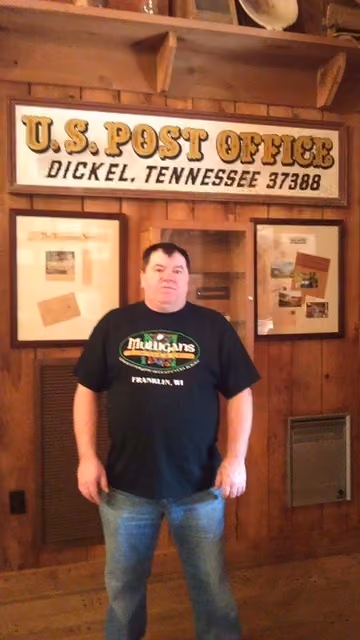 Man in black T-shirt and jeans standing indoors under a sign that says U.S. Post Office Dickel, Tennessee.