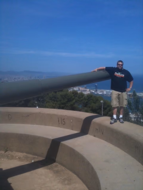 Man standing on concrete platform next to a large artillery cannon overlooking a coastal city and sea.