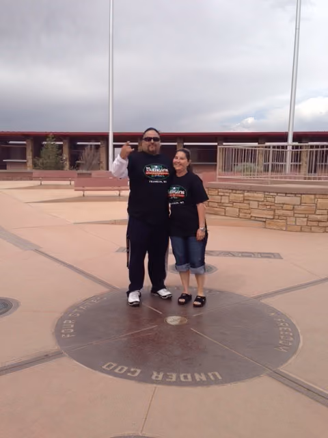 Two people wearing Mulligan's Irish Pub shirts stand on the Four Corners marker outdoors.