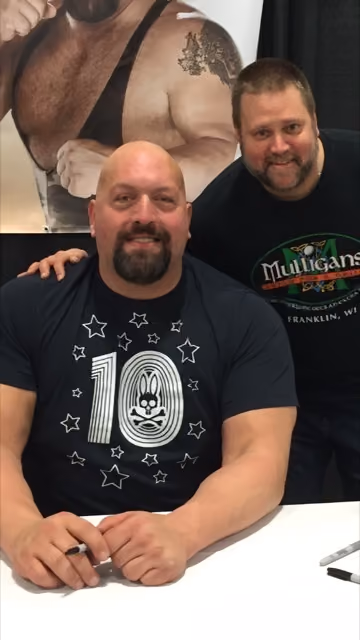 Two men smiling, one seated with a pen, the other standing behind him in a Mulligans pub shirt.