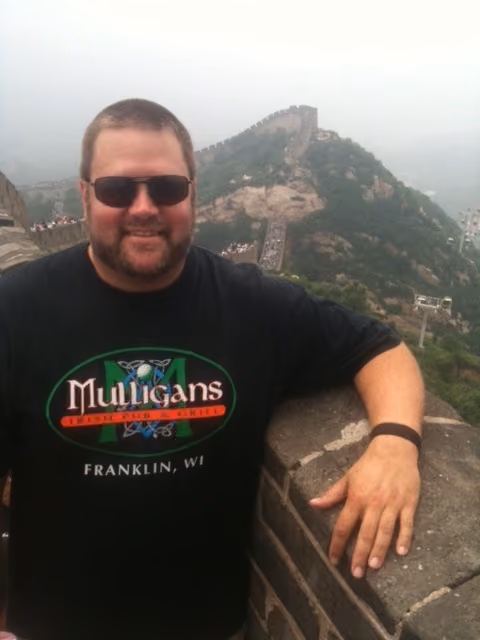 Man wearing sunglasses and a Mulligans Irish Pub t-shirt standing on the Great Wall of China.