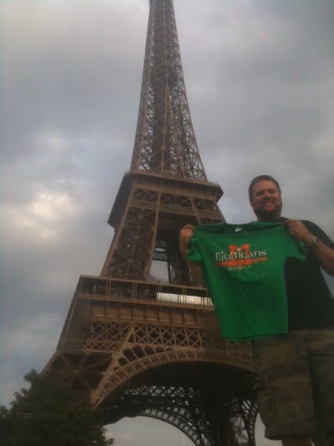 Man smiling, holding a green Mulligans Irish Pub shirt near the base of the Eiffel Tower on a cloudy day.