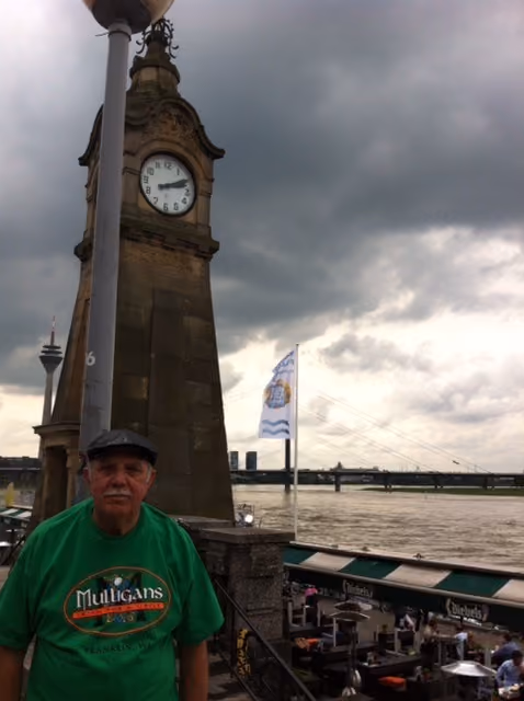 Man in green Mulligans t-shirt standing by a clock tower near a river with cloudy skies overhead.