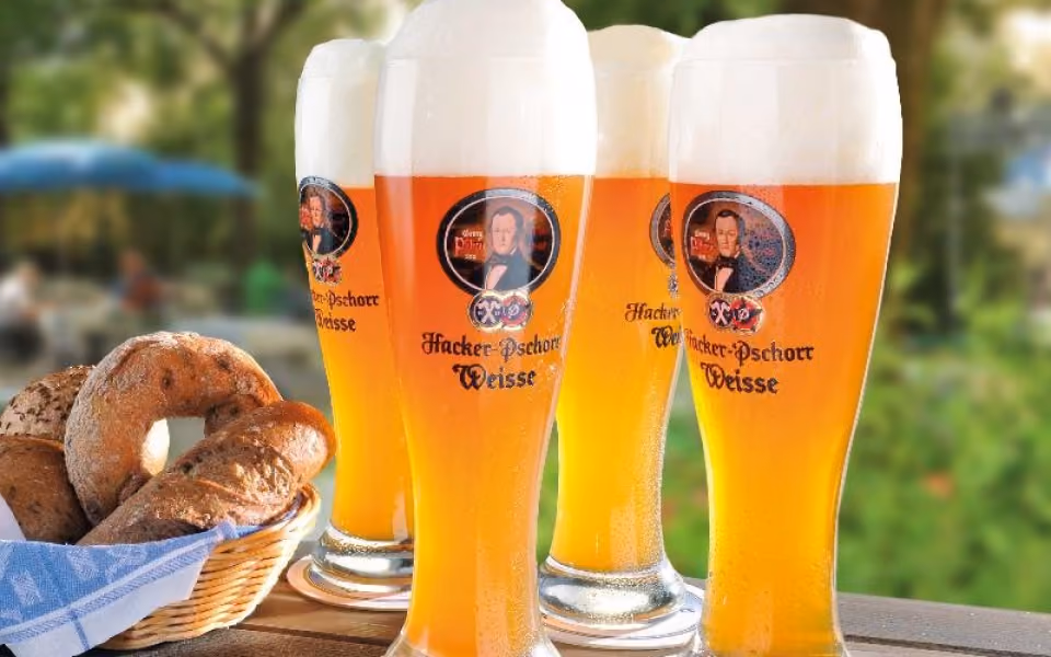 Four tall glasses of Hacker-Pschorr Weisse beer with frothy heads on a wooden table next to a basket of bread.