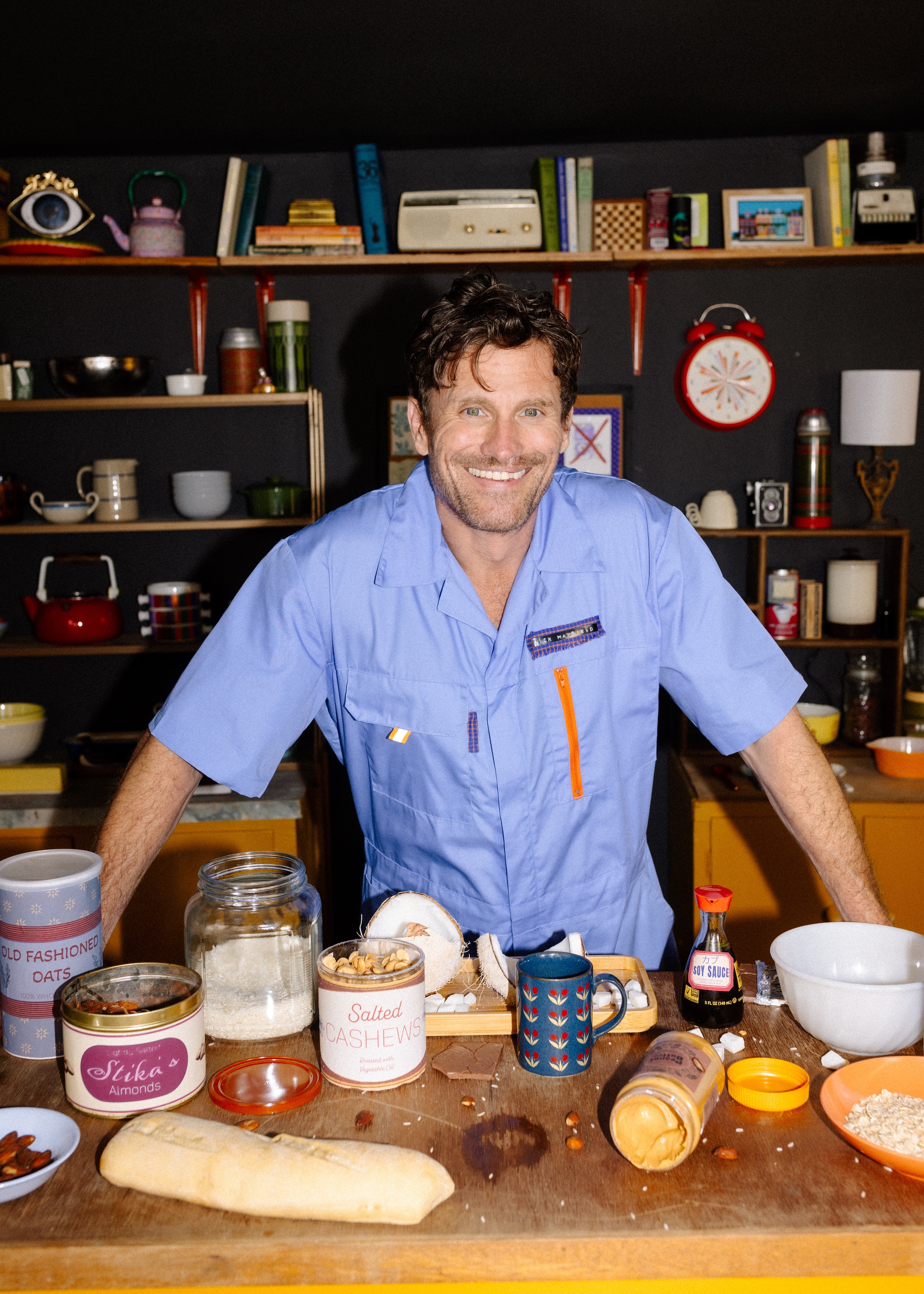 Smiling man in blue shirt stands behind table with oats, salted cashews, peanut butter, soy sauce, and bread.