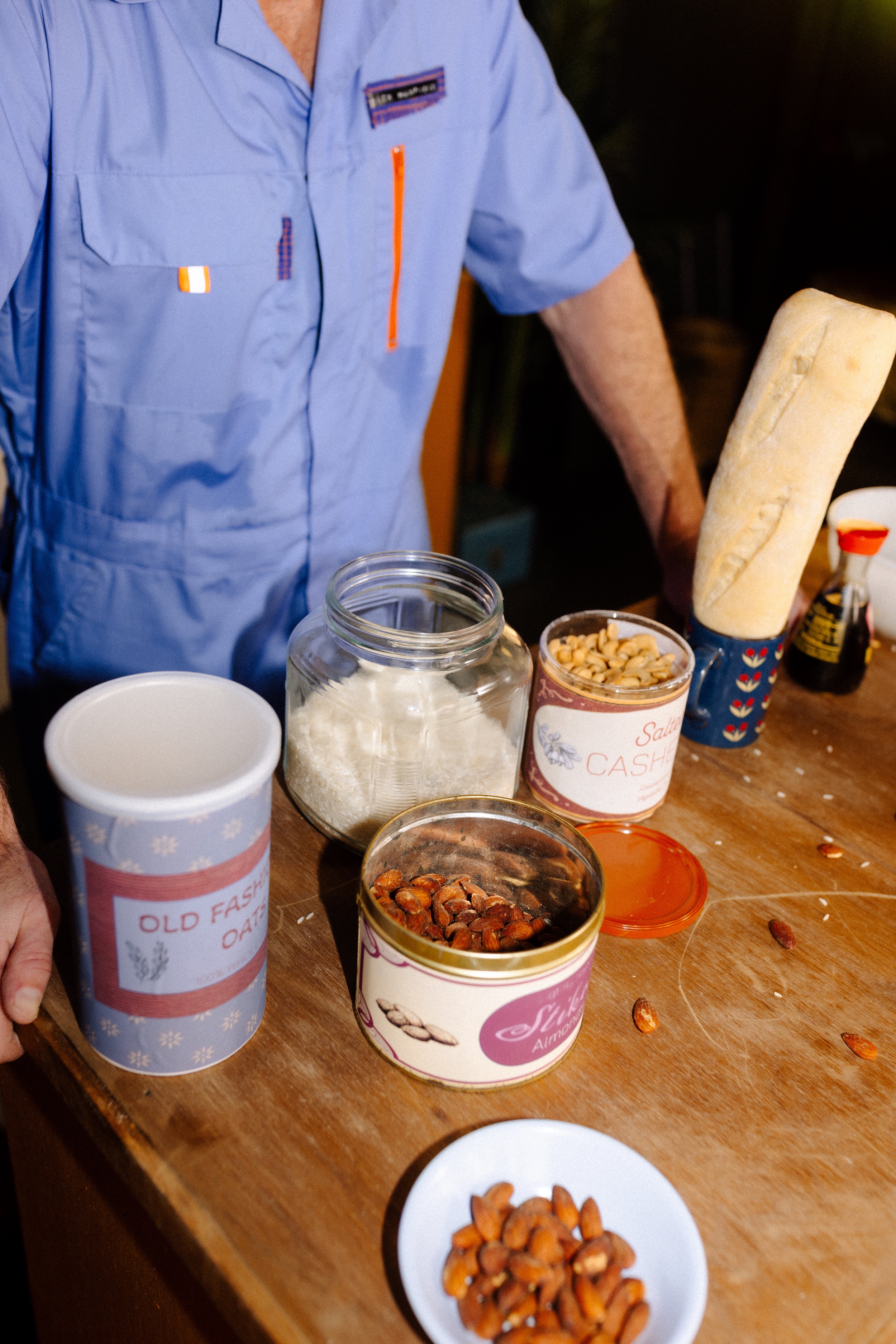 Person in blue shirt stands by a table with containers of oats, rice, cashews, almonds, and a baguette.