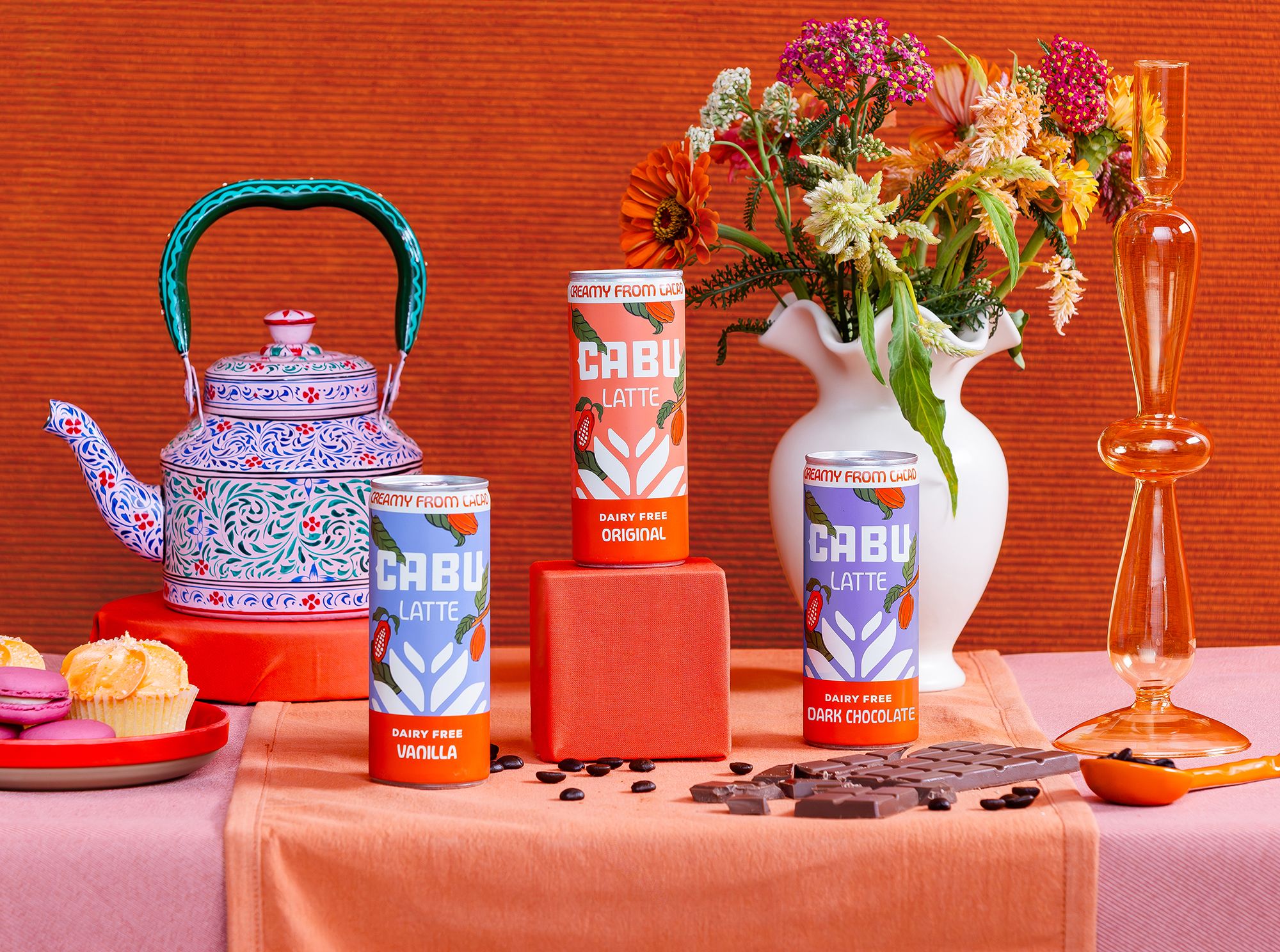 Three Cabu Latte cans with vanilla, original, and dark chocolate flavors on orange and pink cloth.