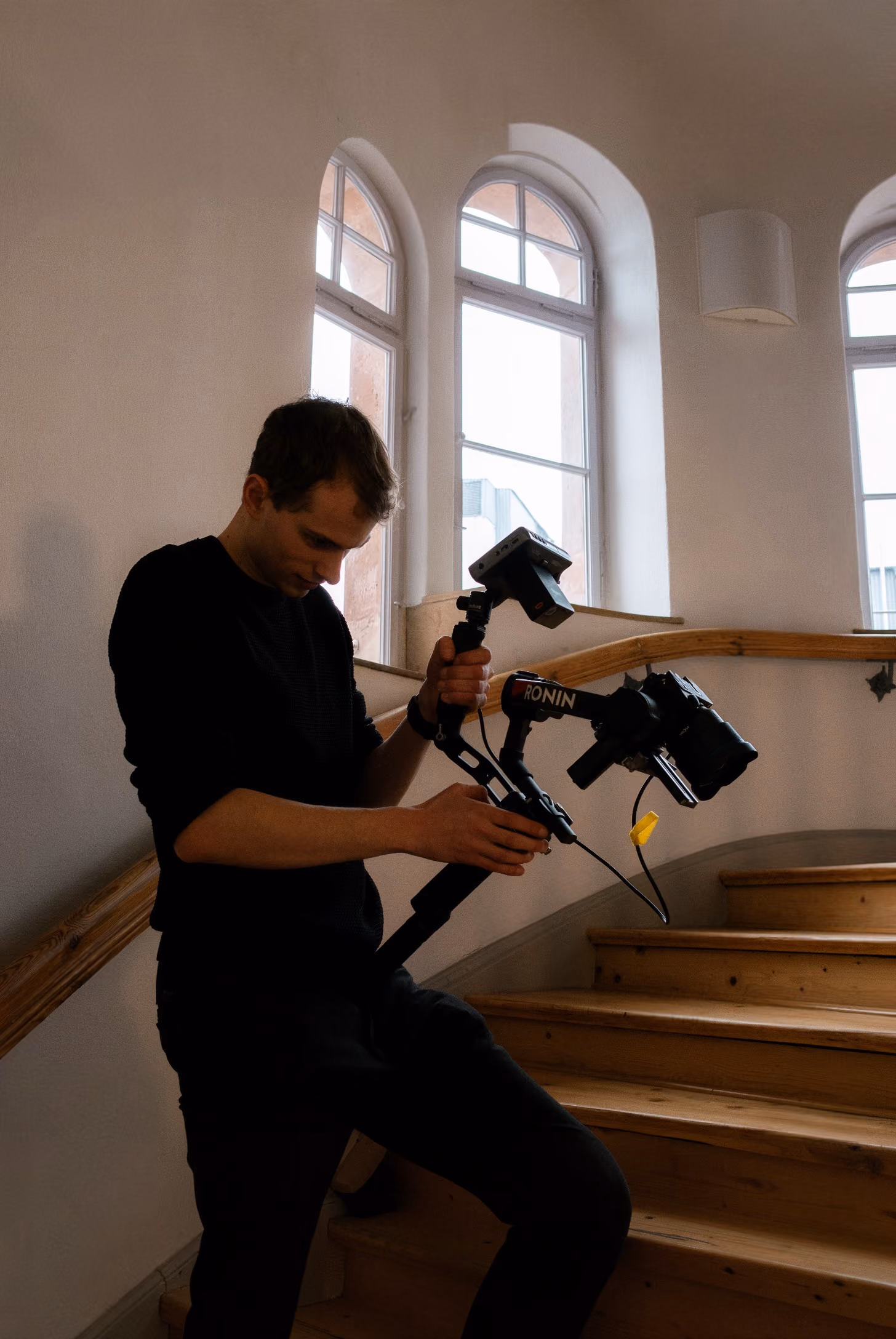 Man adjusting camera mounted on Ronin stabilizer while standing on wooden stairs indoors.