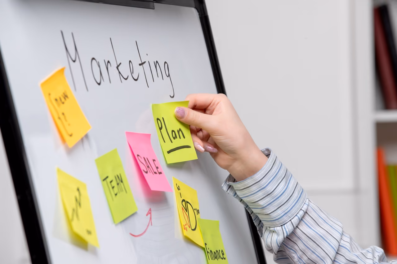Hand placing a sticky note labeled 'Plan' on a whiteboard with marketing strategy notes.