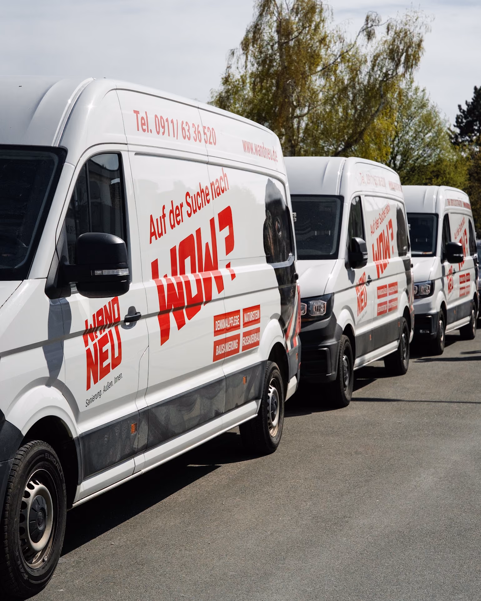 Three white vans with red German text parked on a road near trees in sunlight.