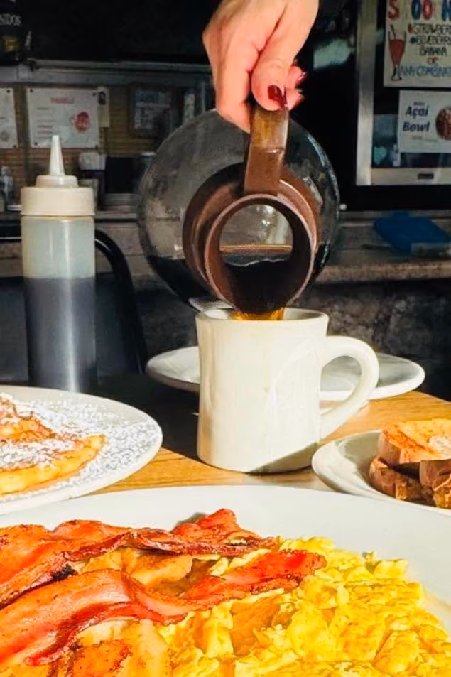 Hand pouring coffee from pot into white mug with bacon, scrambled eggs, and toast on plates.