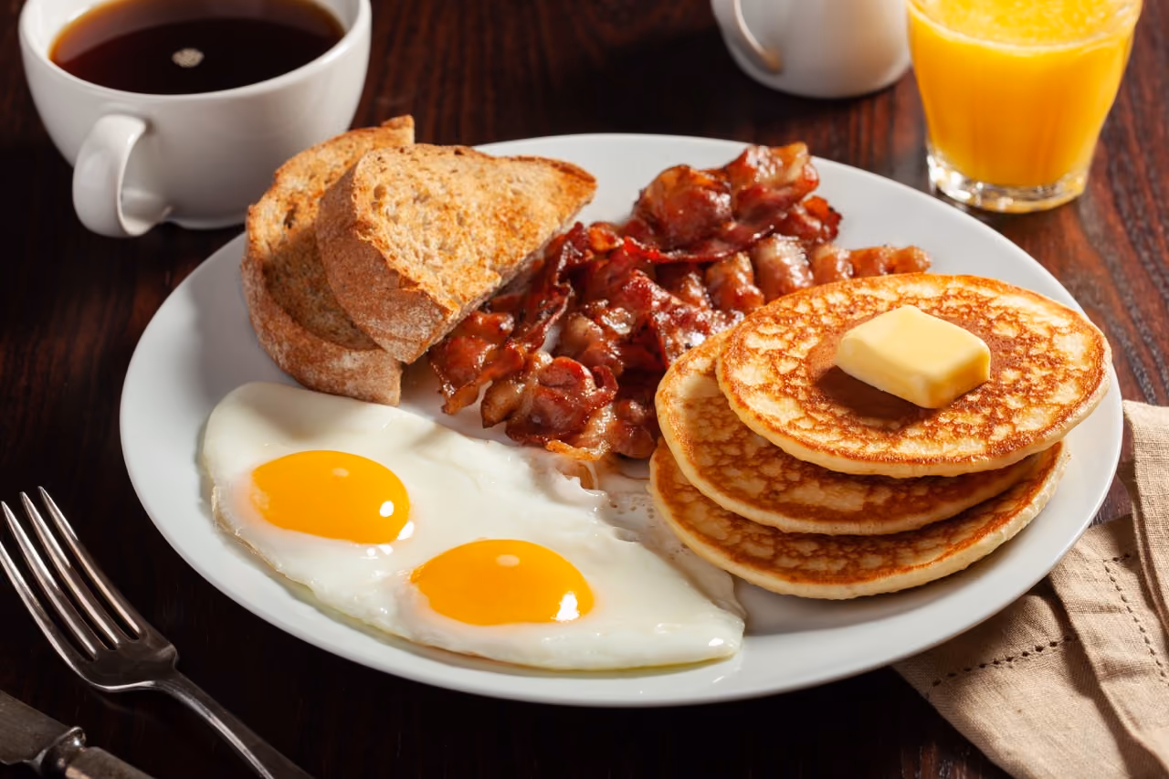 Plate with two fried eggs, crispy bacon, three pancakes topped with butter, and toast with coffee and orange juice