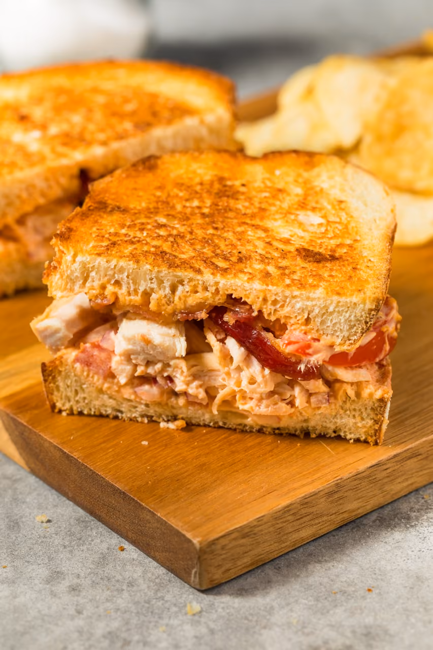 Grilled chicken sandwich with tomato and bacon on toasted bread on a wooden board.