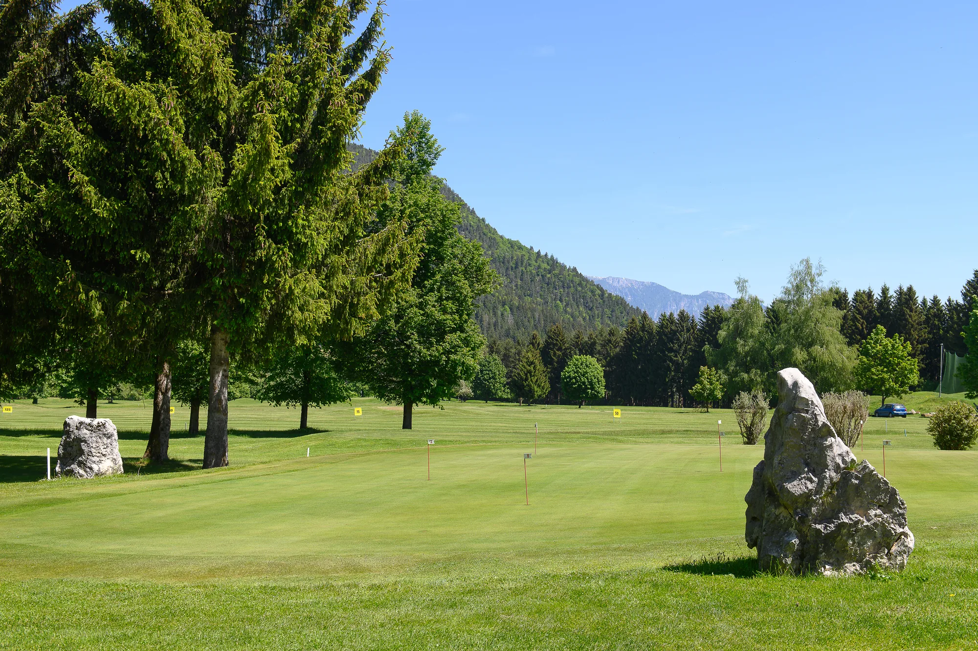 Übungsgrün mit Fahnen und Findlingen vor den bewaldeten Bergen in Tarvisio
