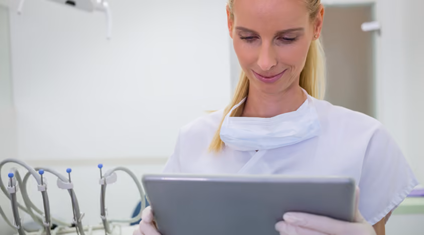 Female dentist with mask and gloves looking at a tablet in a dental clinic.