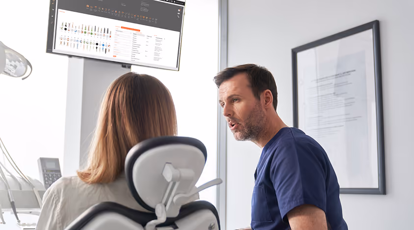 A male dentist in scrubs talking to a female patient sitting in a dental chair with dental tools nearby.