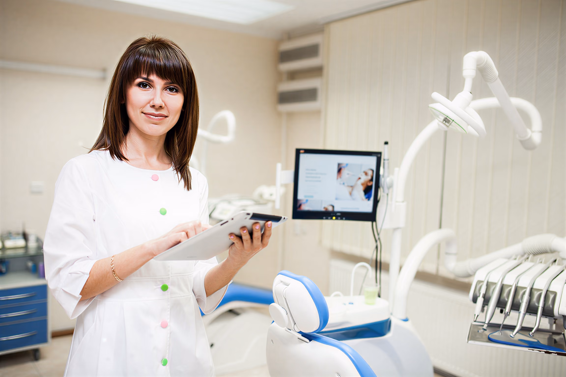 Female dentist in white coat holding tablet in a modern dental office with chair and equipment.