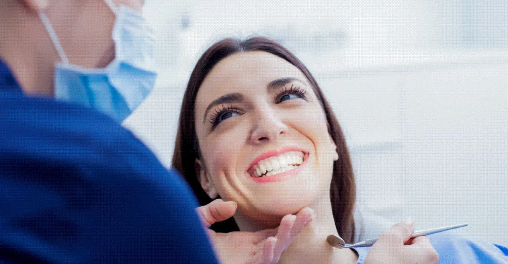 Smiling woman with white teeth at dental checkup by masked dentist in blue scrubs.