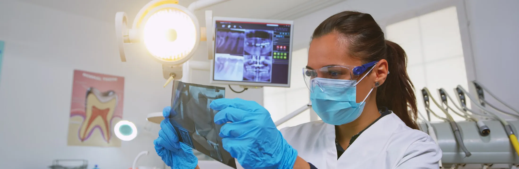 Dentist wearing mask and gloves examining a dental X-ray in a clinic with dental tools behind.