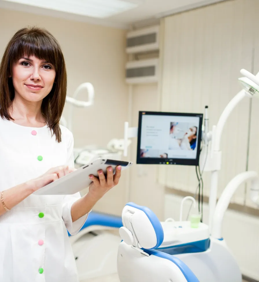 Female dentist in white coat holding a tablet in a dental clinic with dental chair and monitor in background.