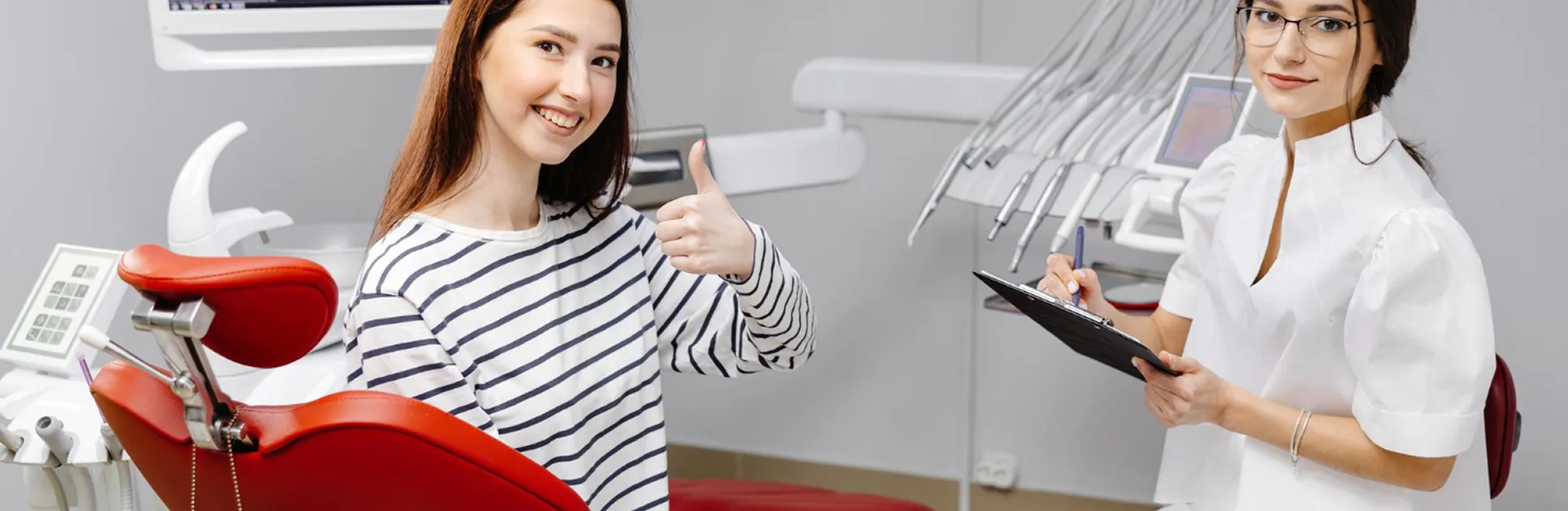 Smiling young woman in a dental chair giving thumbs up to a female dentist with clipboard.
