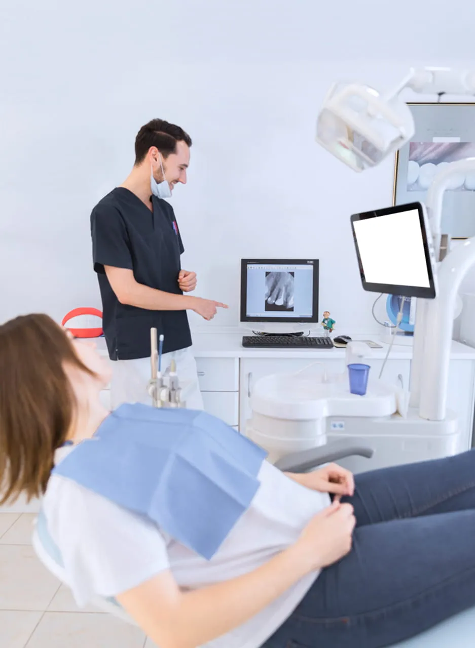 Dentist in scrubs pointing at dental X-ray on computer screen while patient reclines in chair.