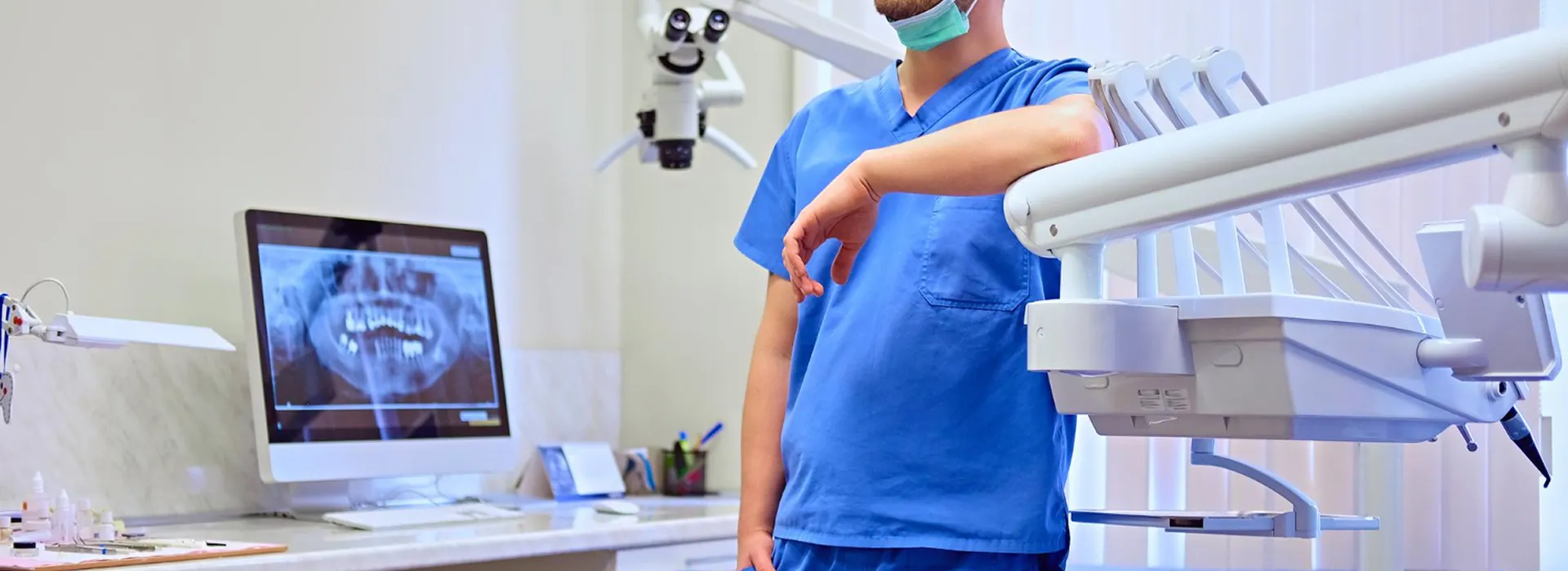 Dentist in blue scrubs and mask standing next to dental equipment and an X-ray image on a monitor.