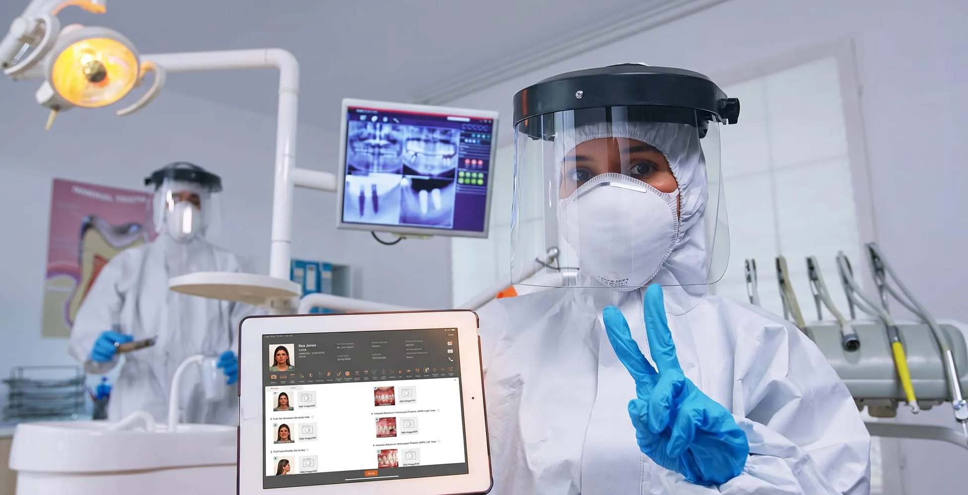 Dental professionals in full protective gear showing dental records on a tablet and making a peace sign.