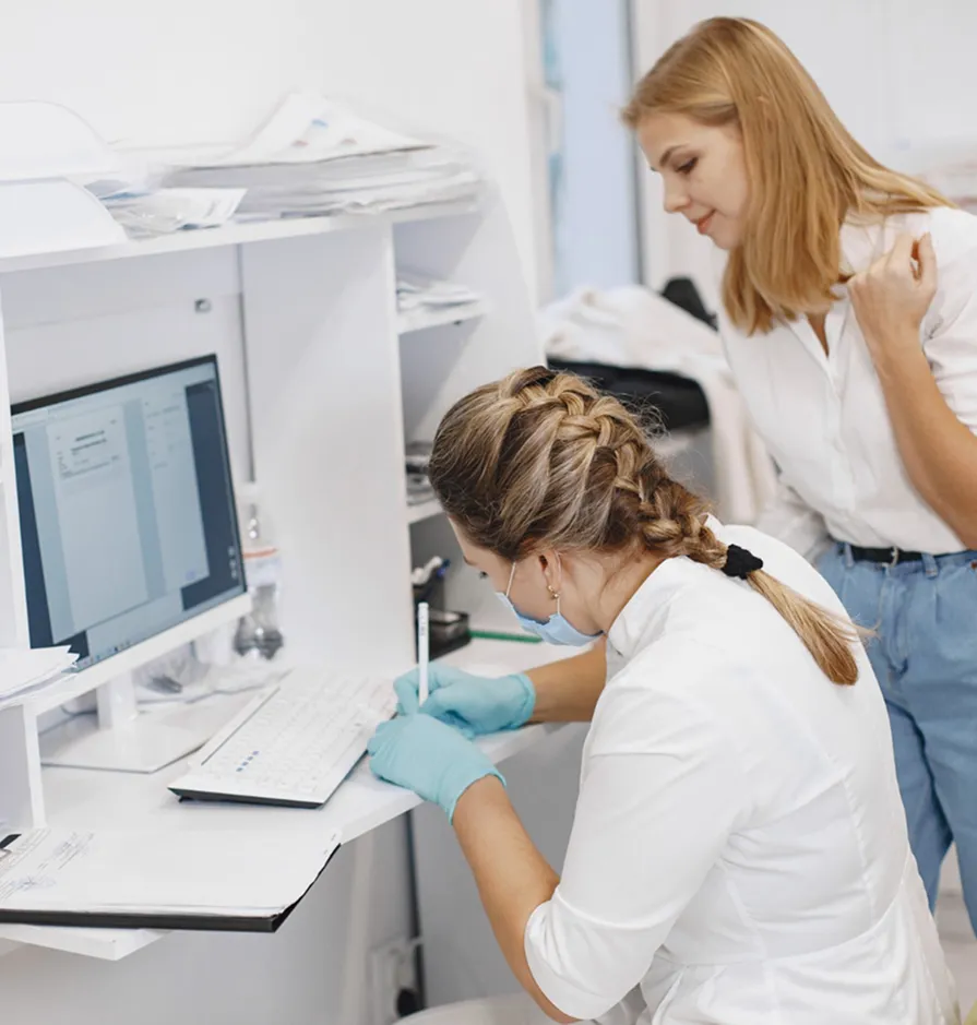 Medical professional in gloves and mask writing notes while colleague watches near a computer monitor.