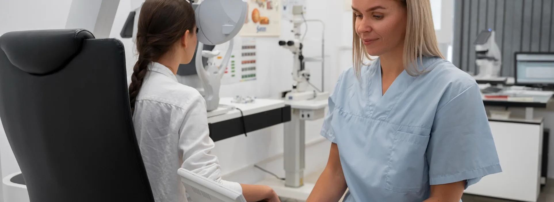 Female eye doctor in blue scrubs examining a young patient with vision test equipment.