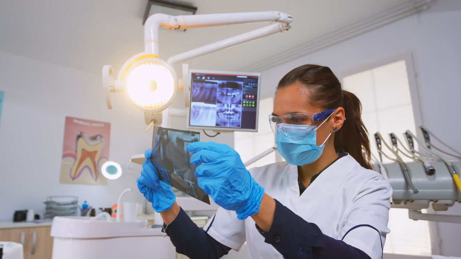Dentist wearing mask and gloves examining dental X-ray film in bright clinic.