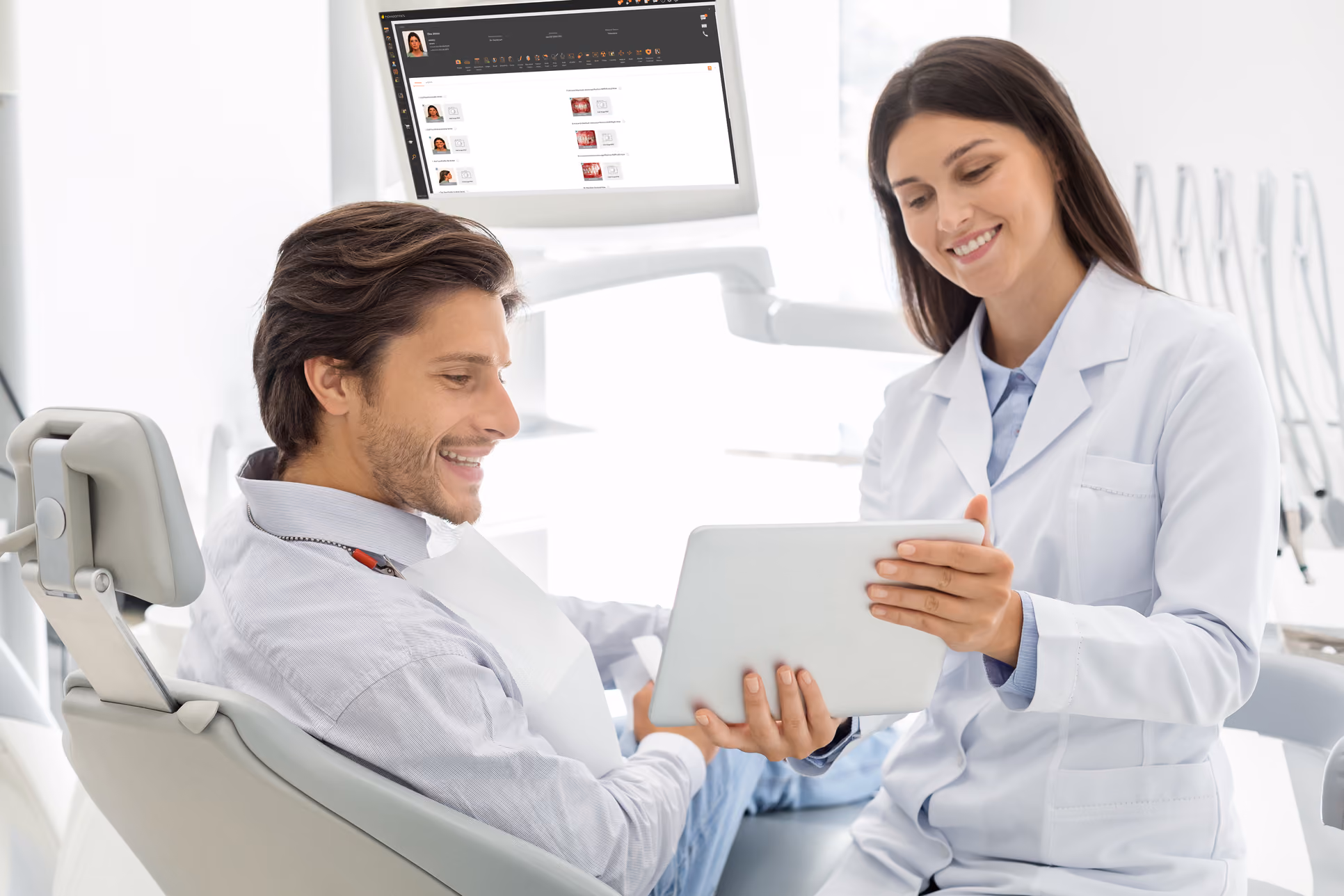 Dentist showing patient information on tablet in a bright dental office with screen display in background.