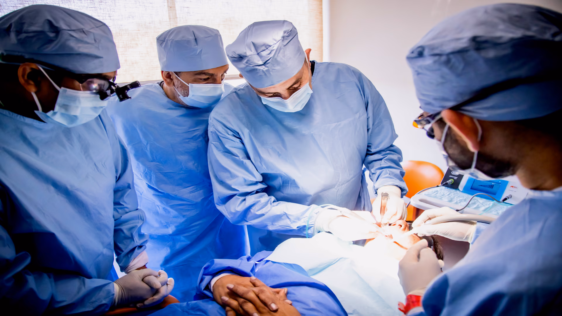 Four surgeons in blue scrubs and masks performing oral surgery on a patient in an operating room.