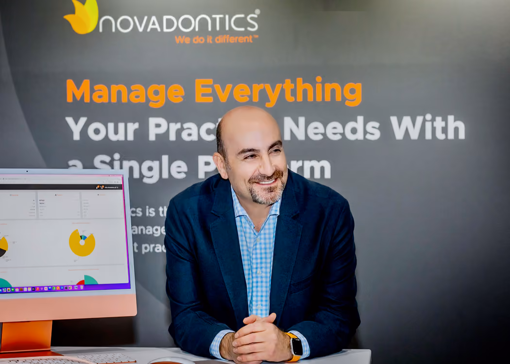Man in blue blazer smiling at desk with computer and Novadontics practice management slogan behind him.