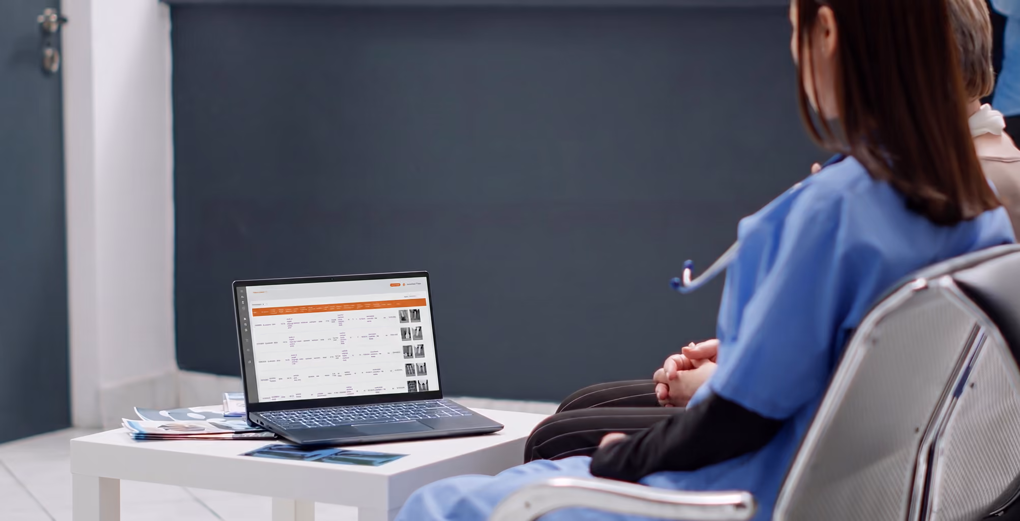 Two healthcare workers in scrubs sitting and looking at a laptop displaying medical data on a table.