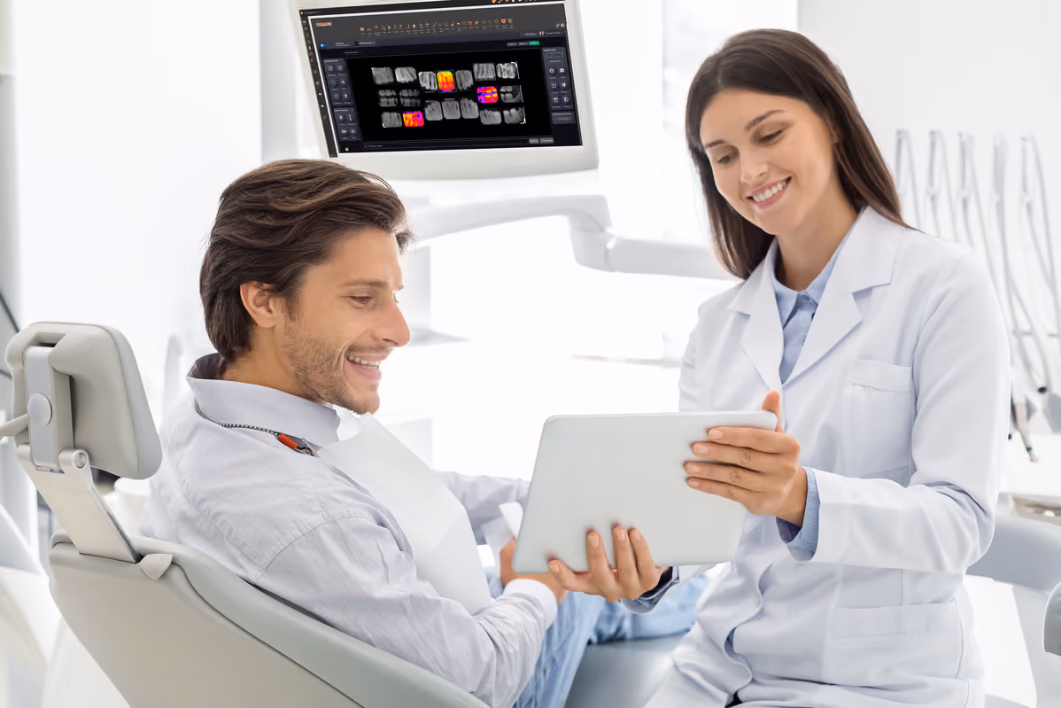 Smiling dentist shows a tablet to a male patient sitting in a dental chair in a bright clinic.