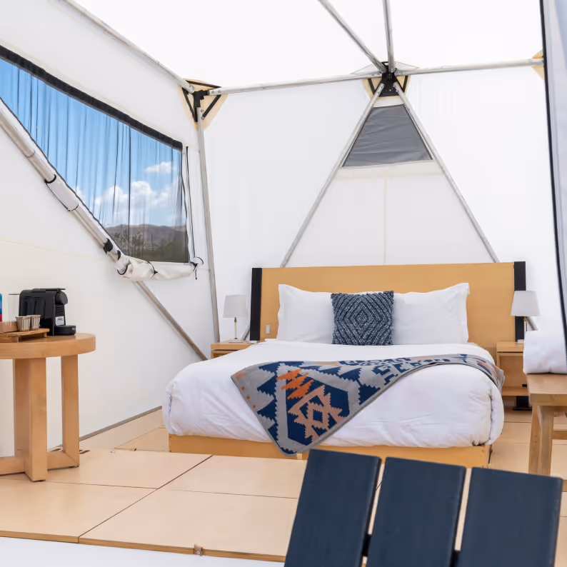 Interior of a bright glamping tent featuring a large bed with white linens, a patterned throw blanket, and a decorative pillow, alongside wooden bedside tables with lamps and a small coffee station.
